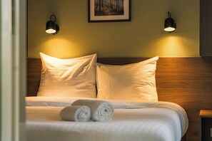 Ein ordentlich bezogenes Bett mit Kissen und aufgerollten Handtüchern, zwei Wandleuchten und ein gerahmtes Bild an der Wand.