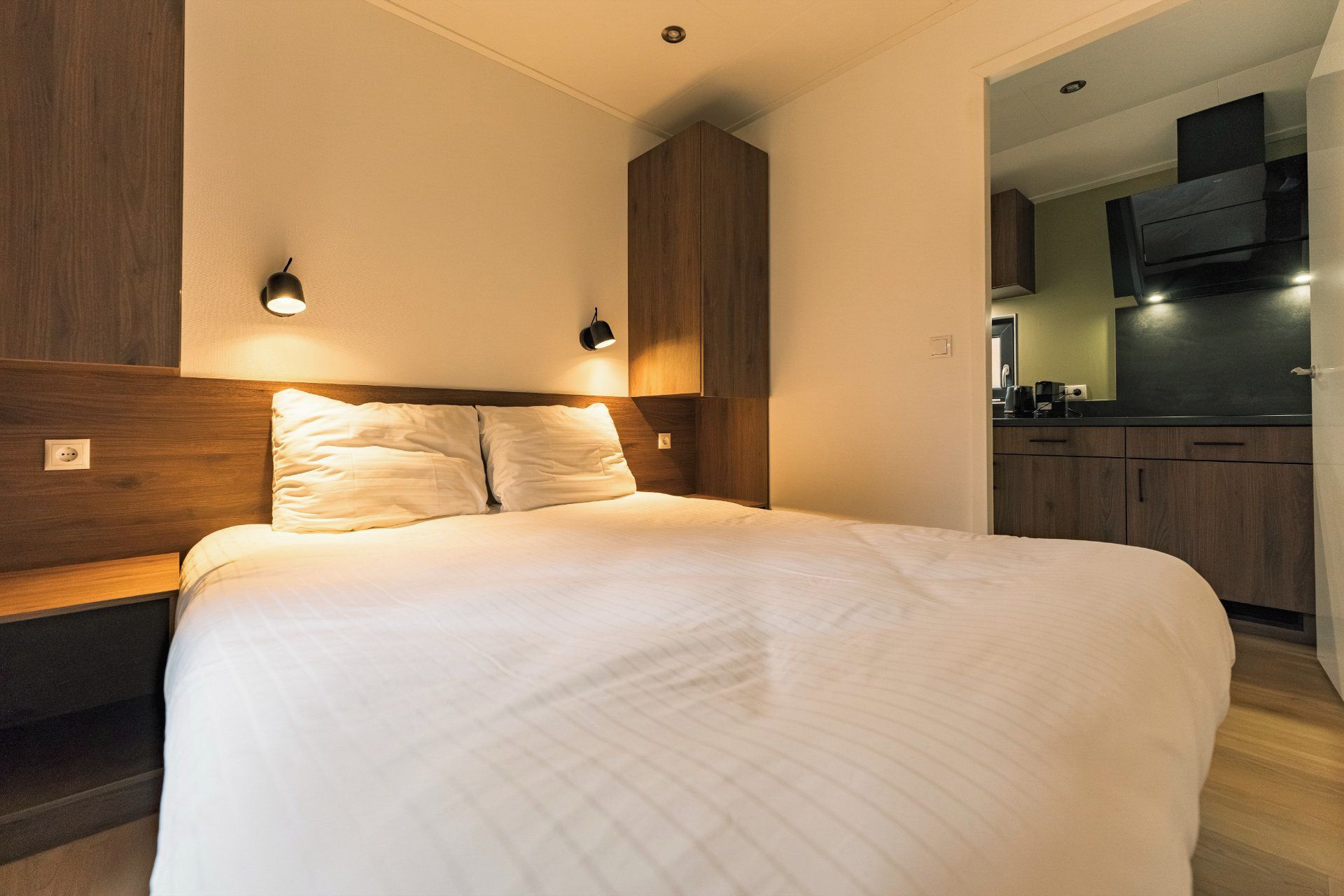 A bedroom with a large bed, wooden headboard, two bedside lamps, and a view into the adjacent kitchen area.