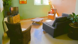 A living room with a television, wicker furniture, a sofa, a table, and a window. - Gite VertCastel – Charming Green Retreat with Pool and Spa (Castelnaudary)
