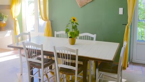 A dining area with a white table and chairs, a hanging pendant light, a wall-mounted basket, and a potted sunflower plant. - Gite VertCastel – Charming Green Retreat with Pool and Spa (Castelnaudary)