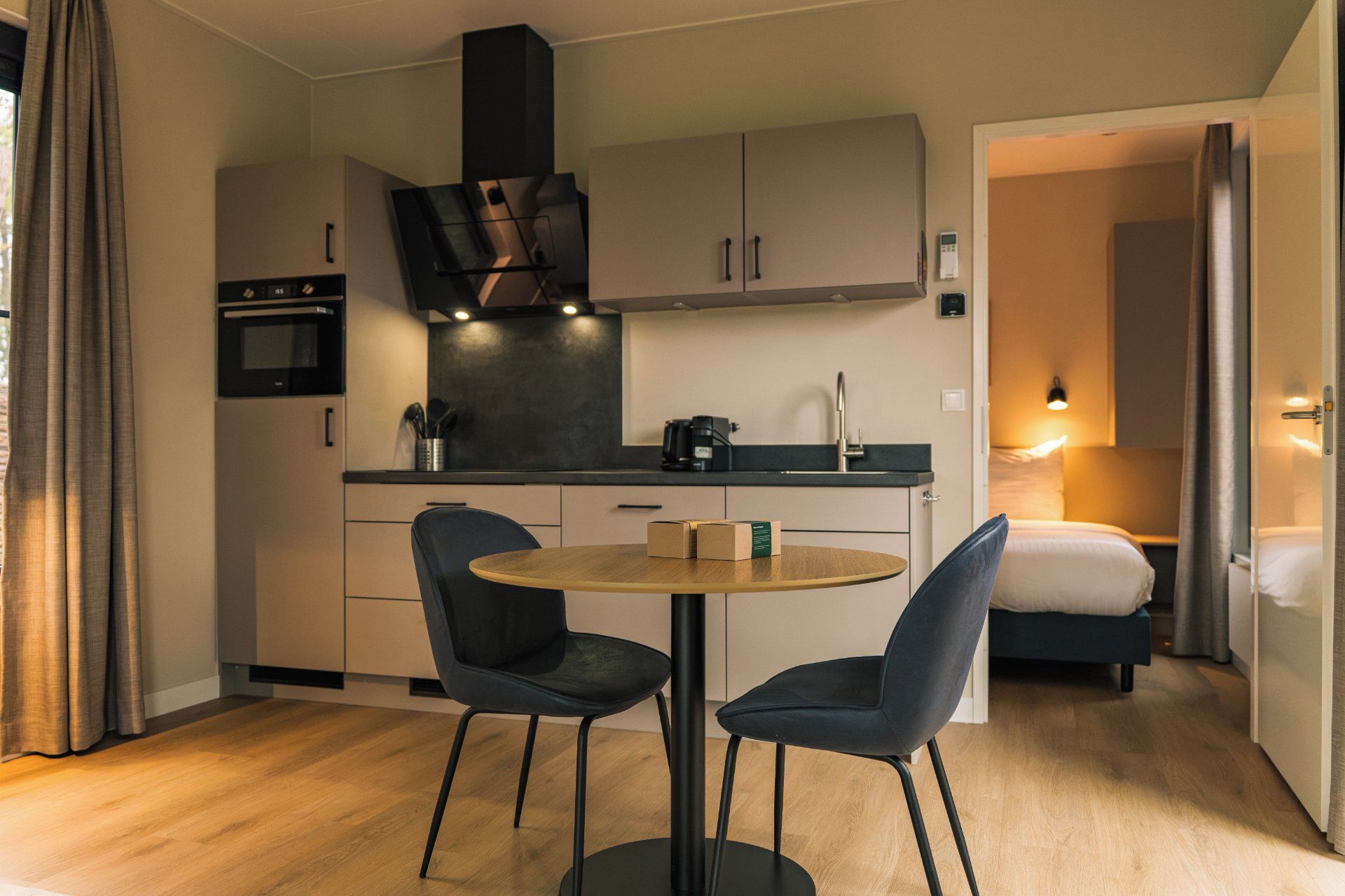 A compact hotel room with a kitchenette, a round dining table with two chairs, a bed, and a bathroom visible through an open door.