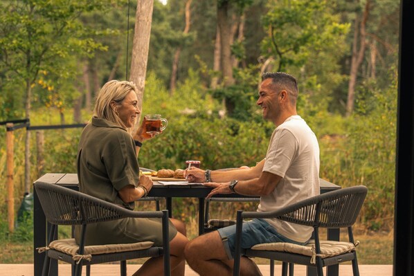 Deux personnes sont assises à une table à l’extérieur, devant des chaises, avec une boisson et une assiette de nourriture.