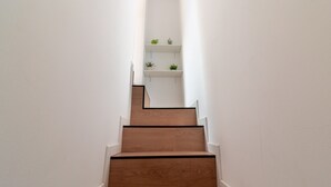 Eine Treppe mit Holzstufen und weißen Wänden, die zu einer Zwischenebene führt, auf der sich zwei Regale mit kleinen Pflanzen befinden.