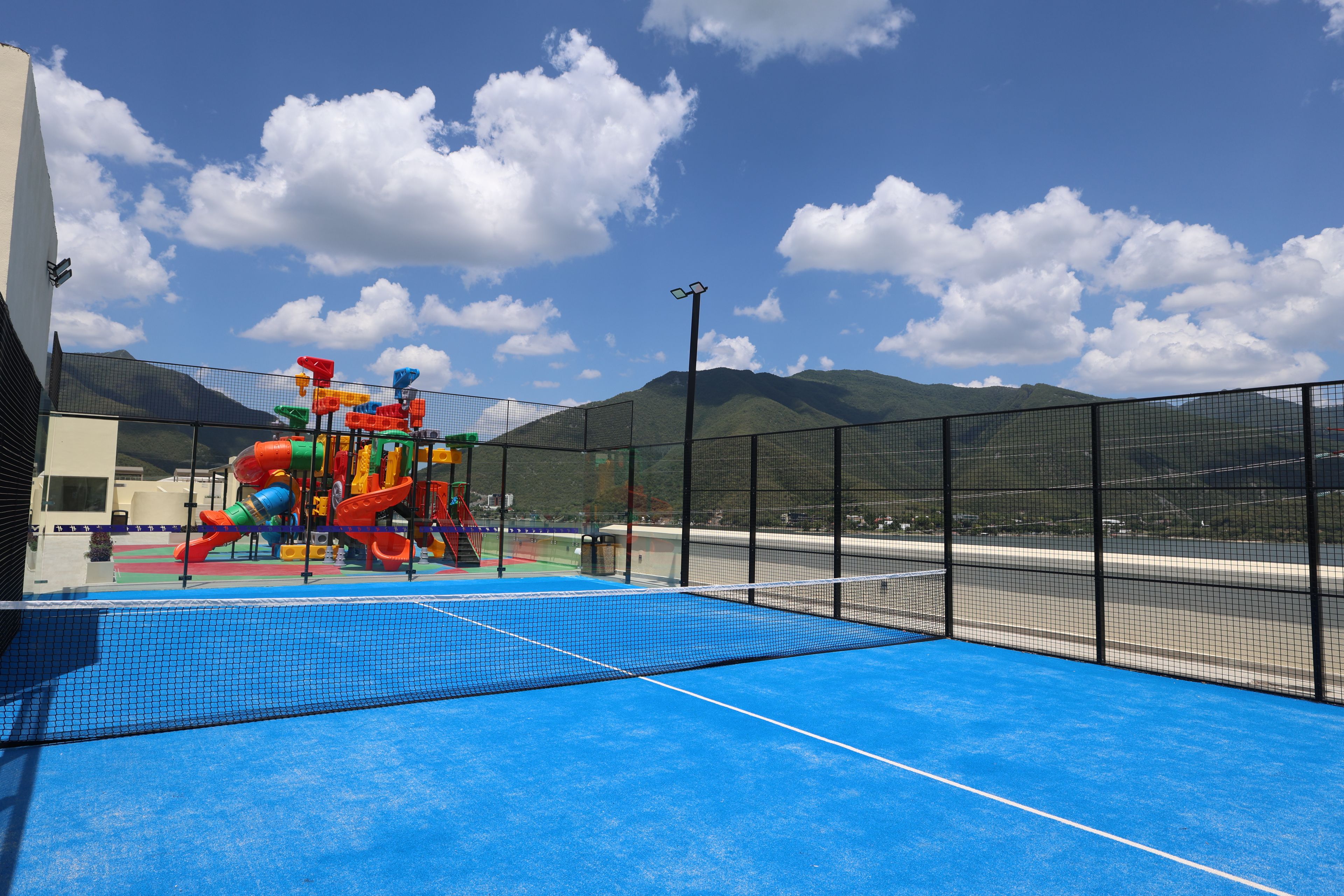 A playground with slides and climbing structures, a blue sports court, and a mountain view in the background.