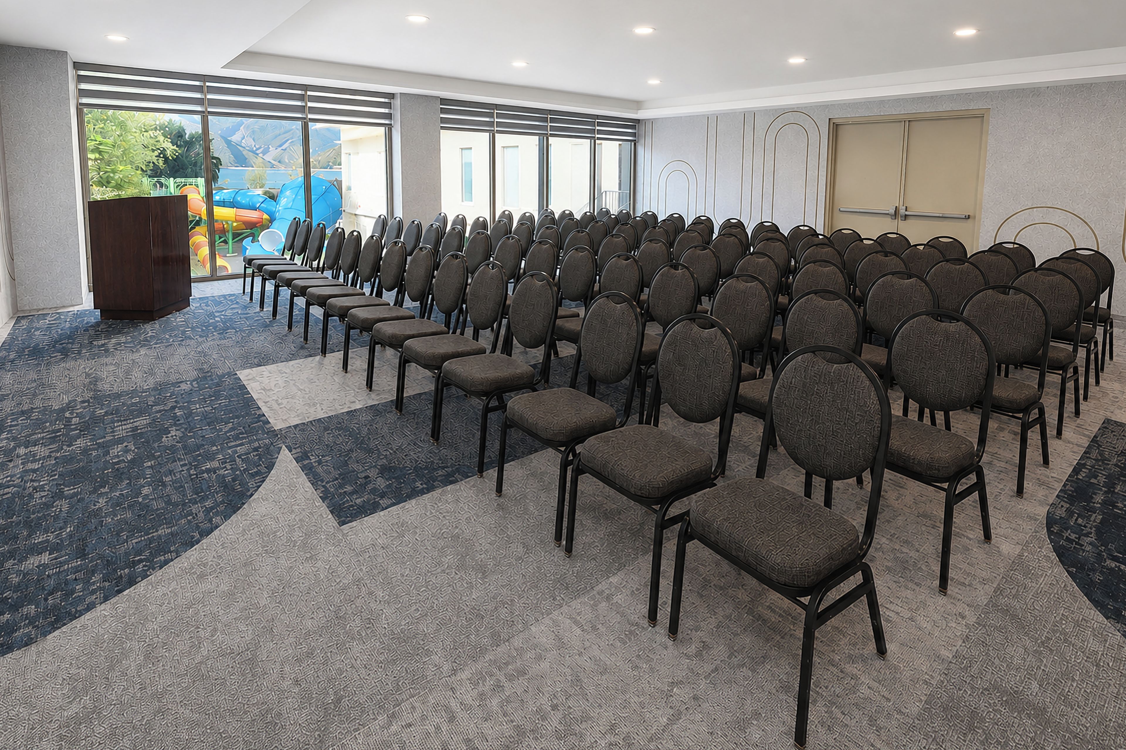 A conference room with rows of chairs, a podium, and large windows with a view of a playground.