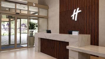 A reception desk with a wooden panel and a marble counter, a large illuminated 'H' sign, and a glass door entrance with a view of the outside.