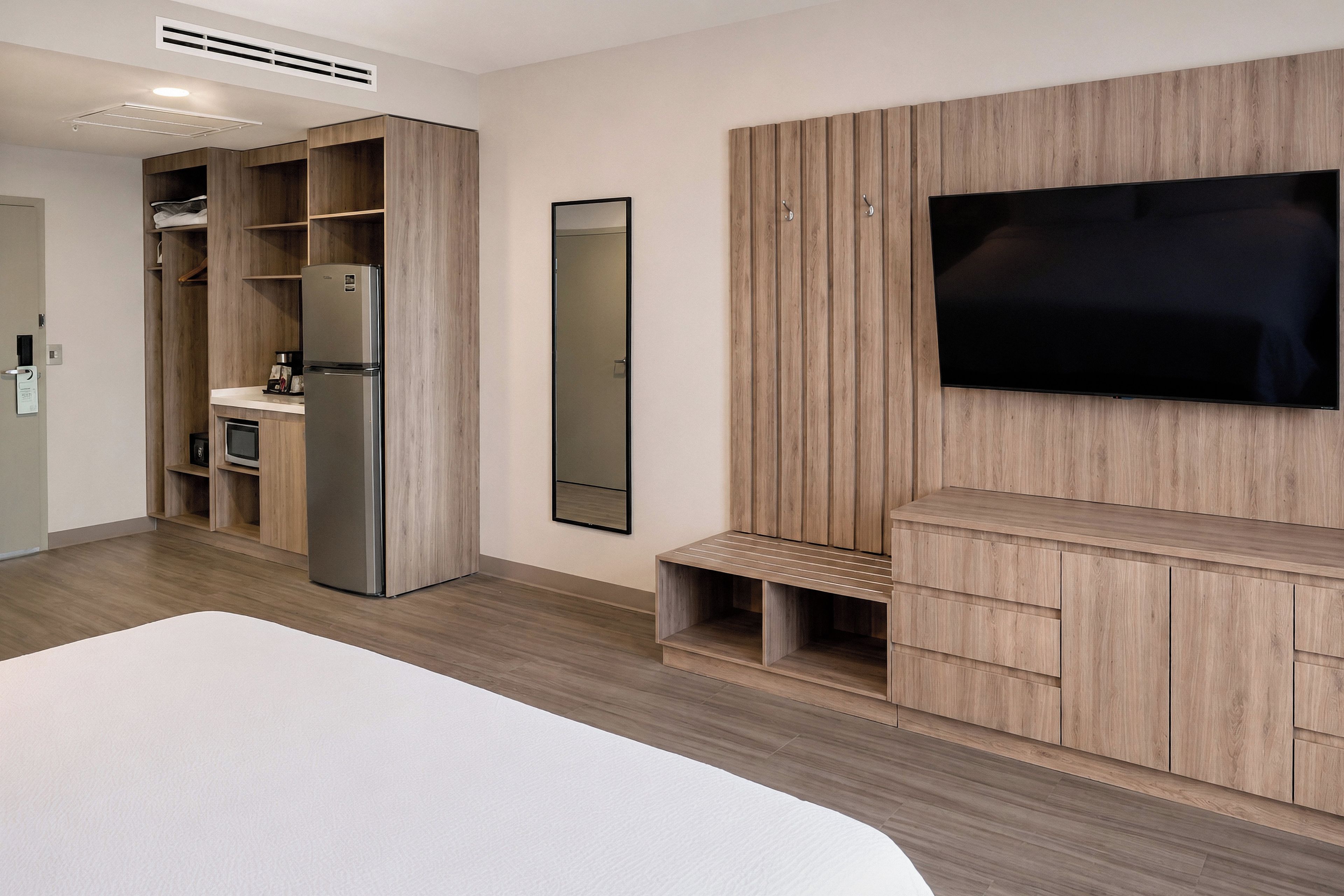 A modern hotel room with a large bed, built-in wardrobe, a kitchen area, and a mounted TV.