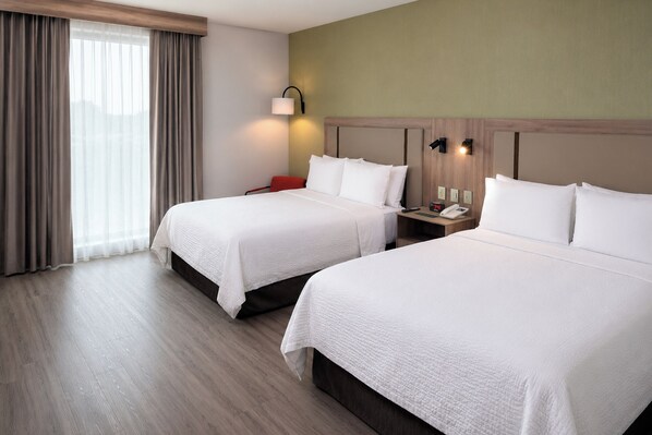 Habitación de hotel con dos camas, una mesita de noche con lámpara y vistas al exterior por la ventana.