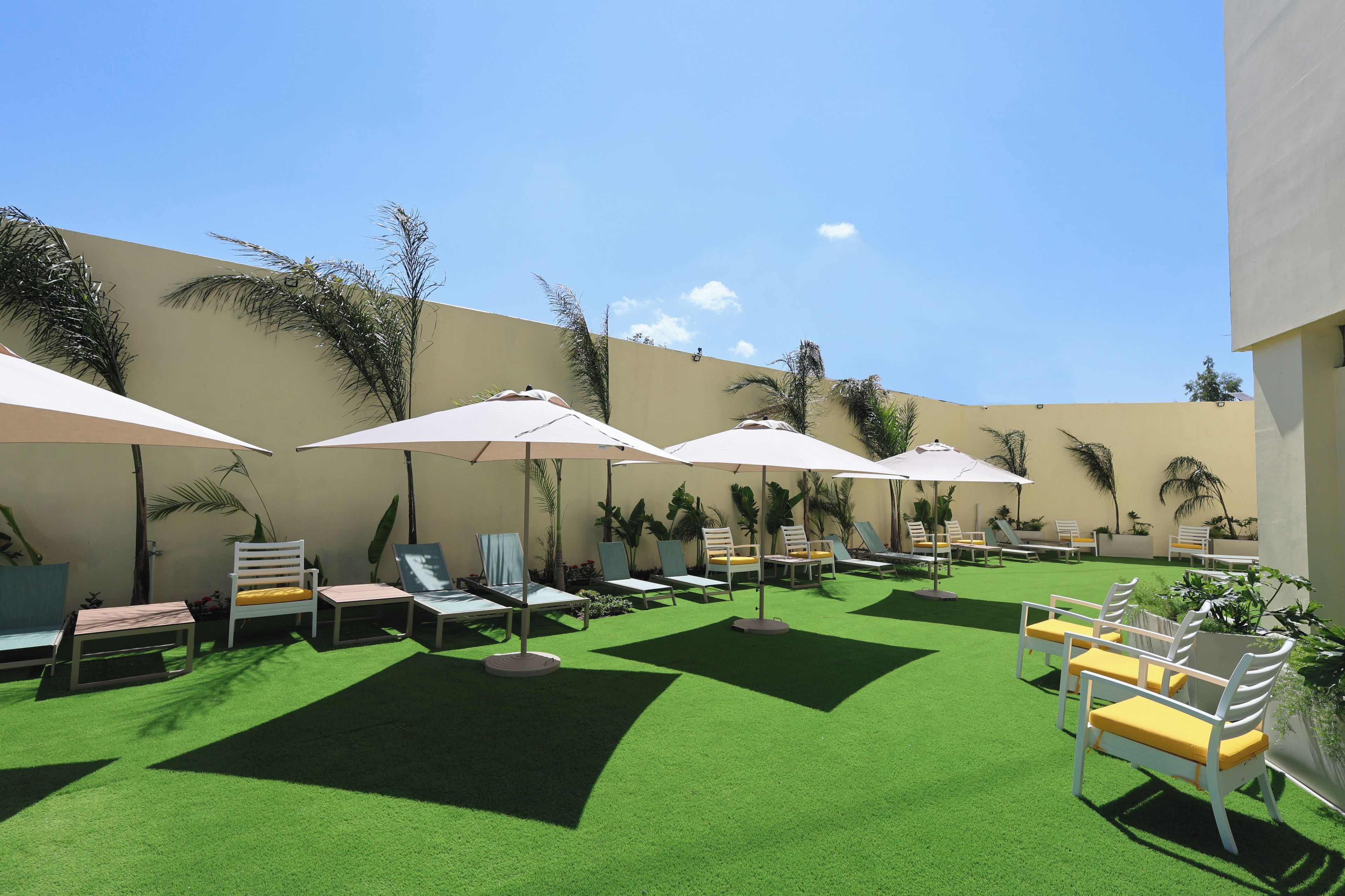 A sunlit outdoor seating area with multiple lounge chairs and umbrellas, surrounded by greenery and a low wall.