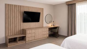 Habitación de hotel con televisión de pantalla plana, escritorio con silla, mesita de noche y ventana grande con cortinas.
