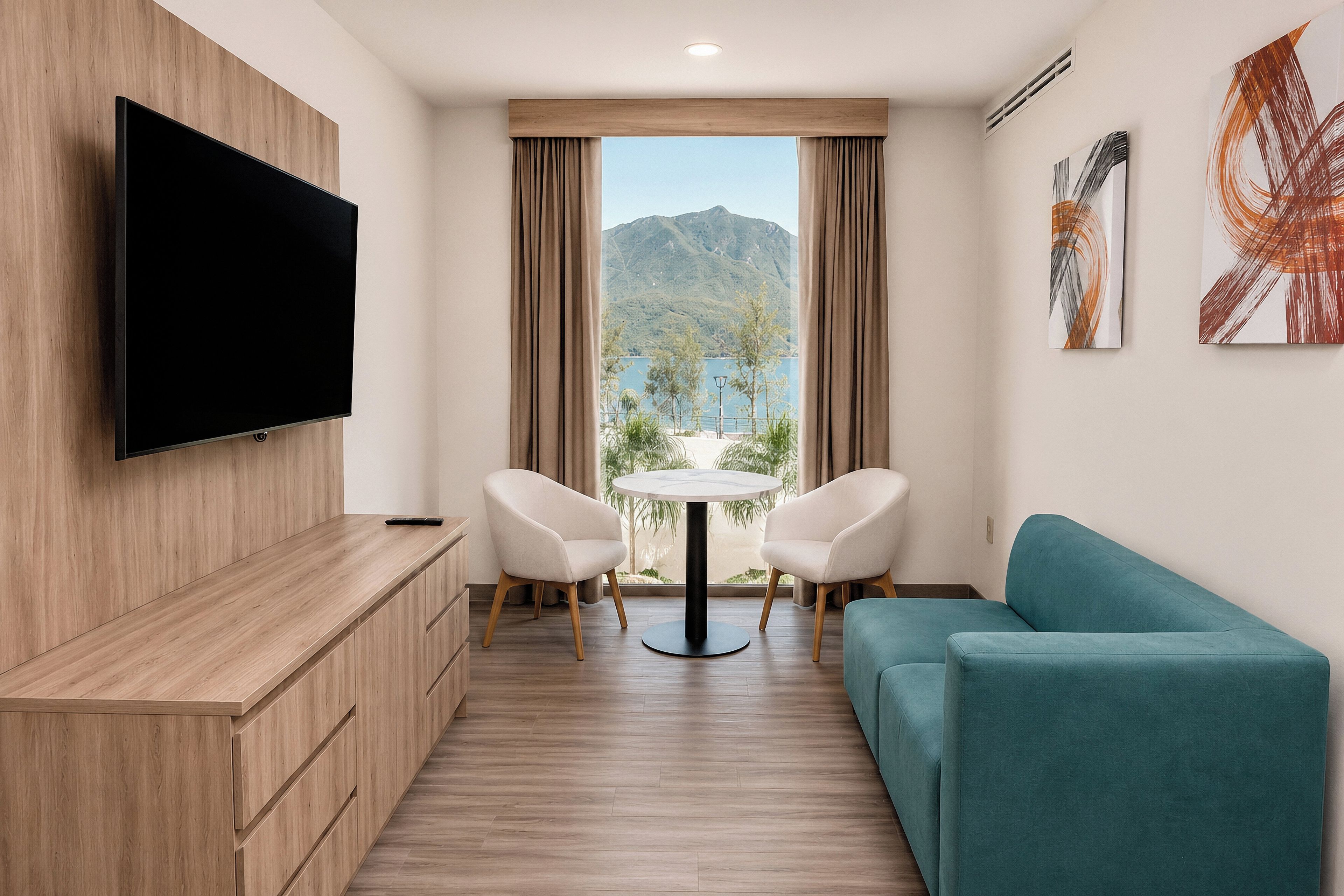 A modern hotel room with a flat-screen TV, a sofa, a dining table with chairs, and a view of mountains through the window.