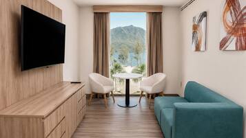 A modern hotel room with a flat-screen TV, a sofa, a dining table with chairs, and a view of mountains through the window.