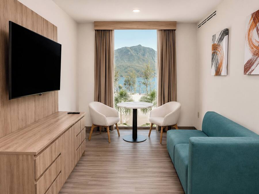A modern hotel room with a flat-screen TV, a sofa, a dining table with chairs, and a view of mountains through the window.