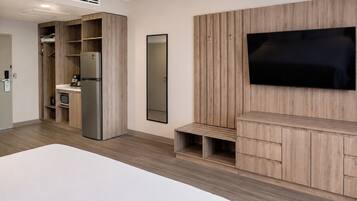 A modern hotel room with a large bed, built-in wardrobe, a kitchen area, and a flat-screen TV mounted on the wall.