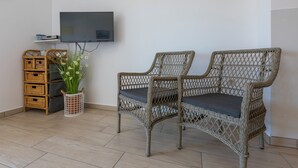 Una sedia in vimini con un cuscino, un mobile contenitore in vimini, un televisore montato a parete e una pianta in vaso con fiori bianchi.