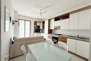Une cuisine moderne avec des meubles blancs, un coin repas doté d’une table en verre et un ventilateur de plafond.