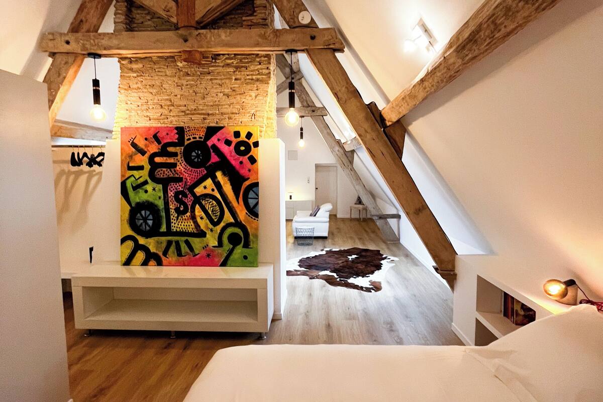 Une chambre mansardée avec des poutres apparentes, une peinture abstraite aux couleurs vives, un tapis en peau de vache et un lit blanc.