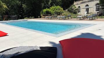 En rektangulär pool med klart, blått vatten, omgiven av ett område med kakelplattor. Det finns solstolar och parasoller, och i bakgrunden syns en byggnad med välvda fönster.