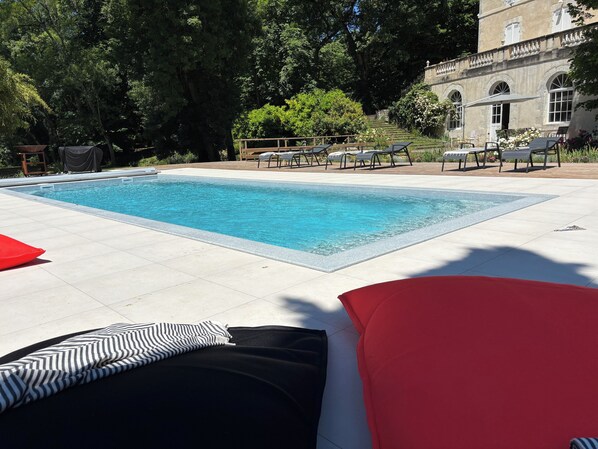 Una piscina rectangular con agua azul cristalina, rodeada de suelo con baldosas. Hay sillas de descanso y sombrillas, y un edificio con ventanas arqueadas al fondo.