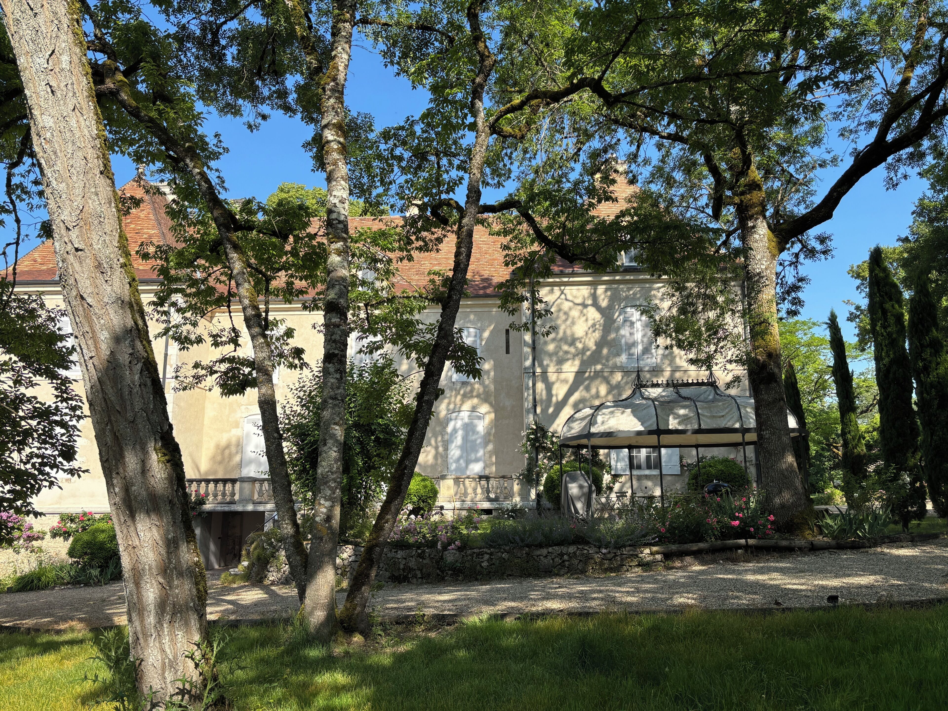 A building with a stone facade, a covered structure with a dome, and a garden with trees and flowers.