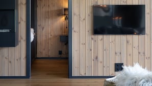 A modern living room with wooden panel walls, a flat-screen TV mounted on the wall, a sofa with a fluffy throw, and a visible door leading to another room. - New (2023) apartment with very central location. (Hol)