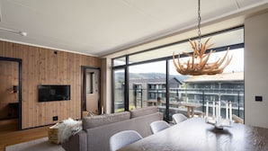 A modern living room with a wooden wall panel, a flat-screen TV, a gray sectional sofa, a dining table with candles, and large windows with a view of buildings and hills. - New (2023) apartment with very central location. (Hol)
