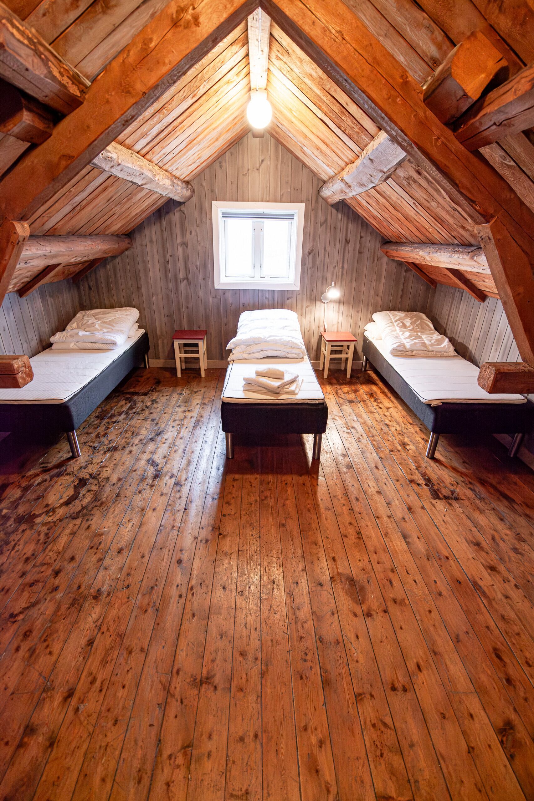 A room with wooden floors, a central bed, two smaller beds, a window, and a light fixture.
