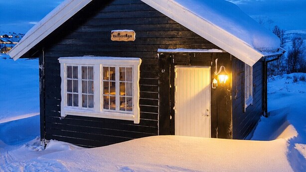 Une cabane en bois, dont le toit est recouvert de neige, avec une entrée illuminée et une enseigne sur laquelle on peut lire « Bidlestugu ».