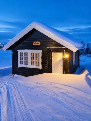 Eine Holzhütte mit schneebedecktem Dach, beleuchtetem Eingang und einem Schild mit der Aufschrift 'Bidlestugu'.