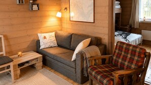 Une pièce chaleureuse avec un plancher en bois, un canapé gris, un fauteuil à carreaux, une petite table avec une lampe et un lit recouvert d’une couverture.