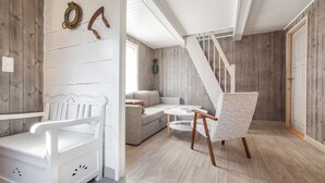 Una habitación con un banco blanco, un sofá, una mesa y una silla. Se observa una escalera y una puerta al fondo.