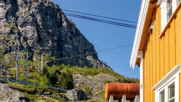 Una terraza de madera con mesas de picnic, un edificio amarillo y una montaña rocosa al fondo.