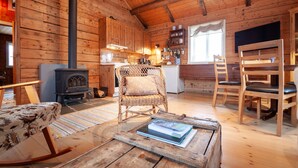 Un intérieur chaleureux de chalet en bois, avec une cheminée, des fauteuils en osier, une table à manger et un coin cuisine.