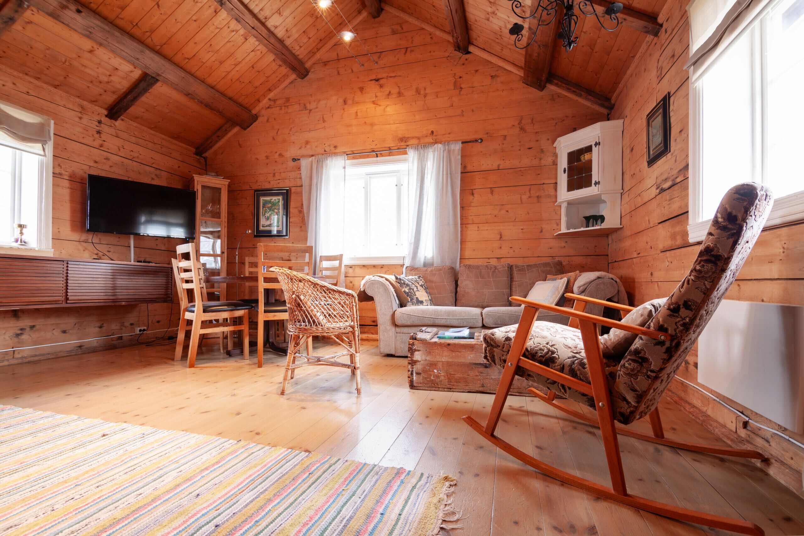 En mysig stuga med träpanel på insidan, ett träbeklätt tak, en tv som hänger på väggen, en soffa, en gungstol och en matplats med stolar och ett bord.