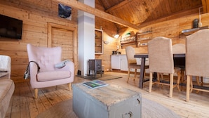 Interior de uma cabana de madeira com sofá, mesa de jantar, cadeiras, pequena cozinha e televisão.