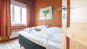 Um quarto com painéis de madeira, uma cama, uma janela com cortinas, um quadro emoldurado na parede e uma porta com puxador.
