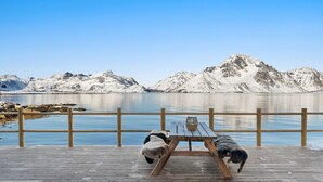 A wooden deck with a picnic table and a basket, overlooking a calm lake with snow-capped mountains in the background.