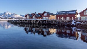 A row of red wooden buildings with large windows, a wooden dock, and a calm body of water reflecting the structures.
