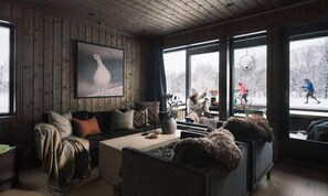 Interior acogedor de una cabaña con techo de madera, un ventanal con vistas a la nieve, una obra de arte enmarcada y una zona de estar confortable.