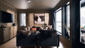 Un soggiorno accogliente con un grande televisore piatto, due divani, pareti rivestite in legno e ampie finestre con vista sull'esterno.