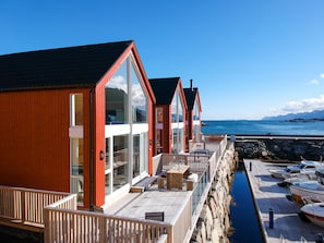 A row of wooden cabins with large windows, a stone wall, and a view of the sea. - High Standard Lodge Ballstad (Vestvågøy)