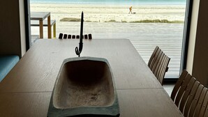 A dining table with a metal sink in the center, wooden chairs, and a view of the beach through a large window.