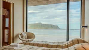 A hotel room with a bed, a chair, and a large window offering a view of a mountain and water.