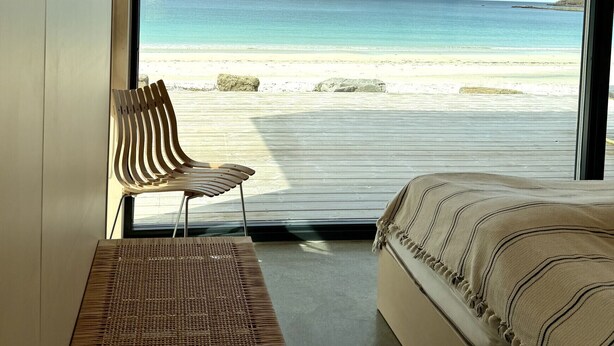A bedroom with a large window overlooking a beach, a bed, a chair, and a woven mat.