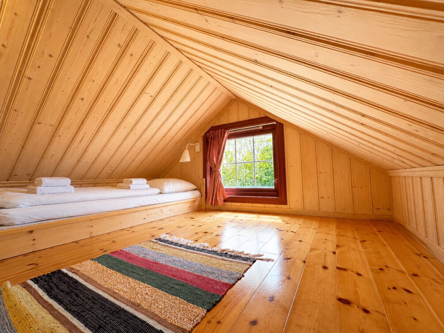 Un dormitorio de ático con piso de madera, una cama, un ventanal y una alfombra colorida.