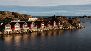 A row of red buildings with white windows and a wooden deck, situated on a rocky shoreline by the water.