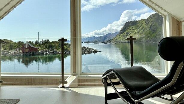 A room with a large window overlooking a fjord, a wooden deck, and a black lounge chair.