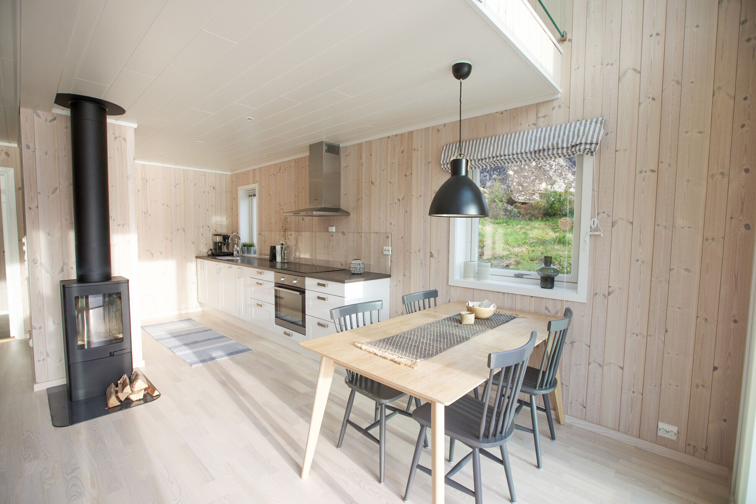A modern kitchen with a wood stove, dining table, chairs, and a hanging pendant light.