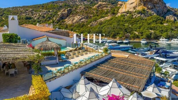 Un hotel sulla costa con piscina sul tetto, montagne sullo sfondo e barche in acqua.