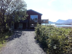 Ein zweistöckiges Gebäude mit Balkon, Kiesweg und Blick auf ein Gewässer und Berge.